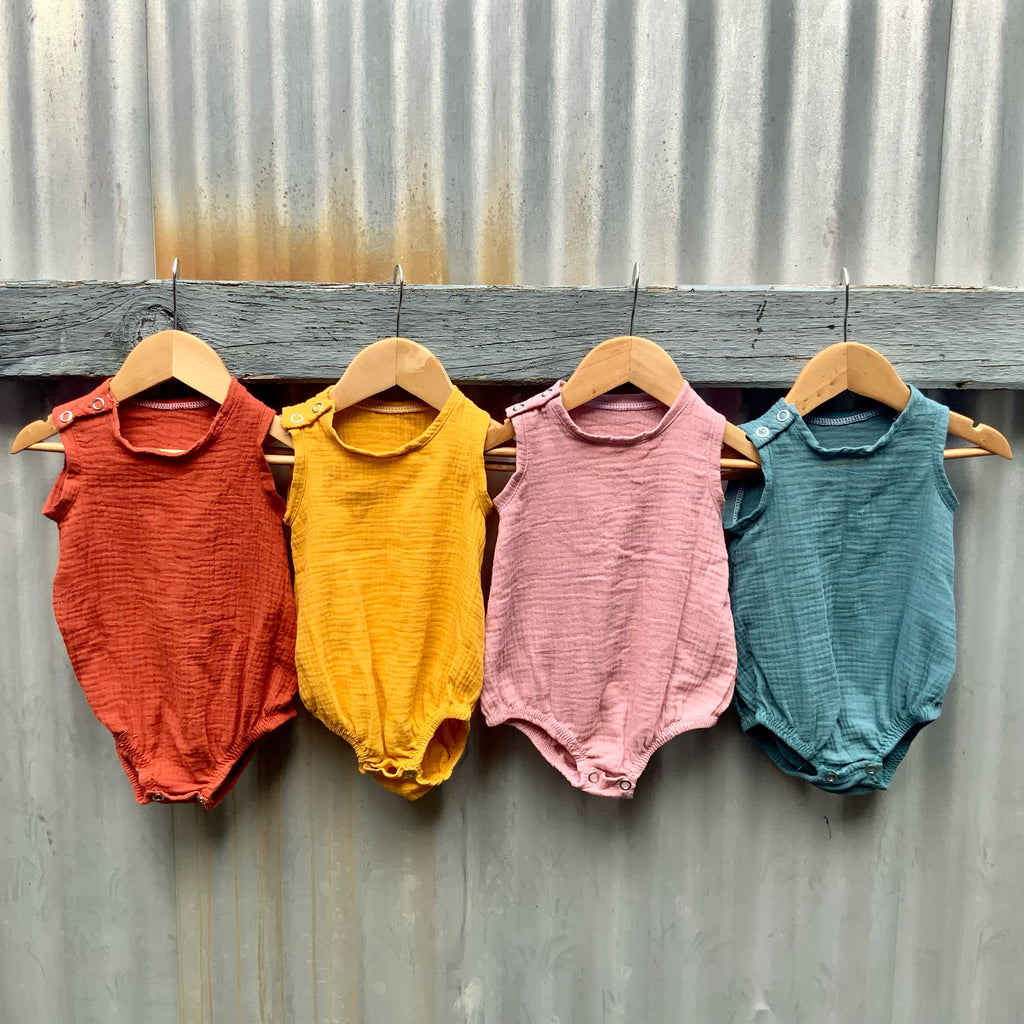Four little Asiki Baby Muslin Cotton Onesies (in red, yellow, pink and green) hanging on a fence.