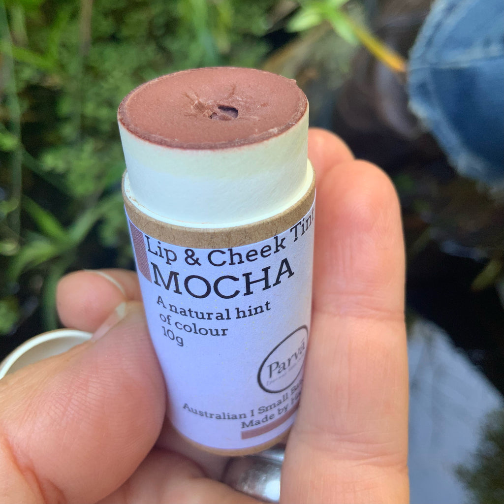 A photo of a hand holding a little cardboard tube of Mocha Lip & Cheek tinted balm by Kirsty Mootz of Parva Little Things organic skincare. Available at Asiki.
