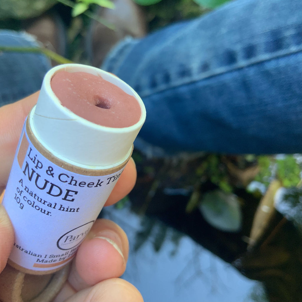 A photo of a hand holding an open little cardboard tube of Nude Lip & Cheek tinted balm by Kirsty Mootz of Parva Little Things organic skincare. Available at Asiki.
